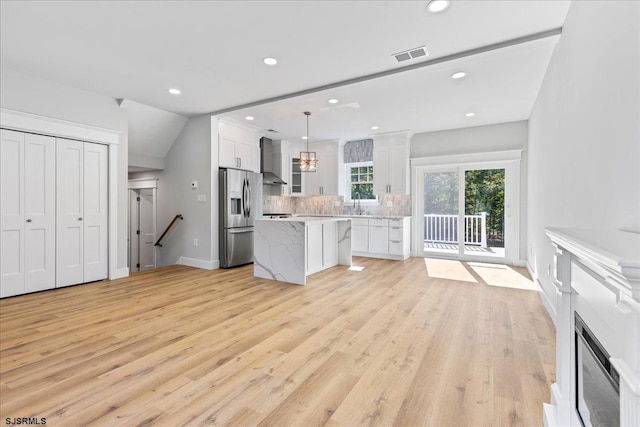 kitchen featuring light wood-type flooring, wall chimney range hood, stainless steel fridge with ice dispenser, white cabinetry, and hanging light fixtures