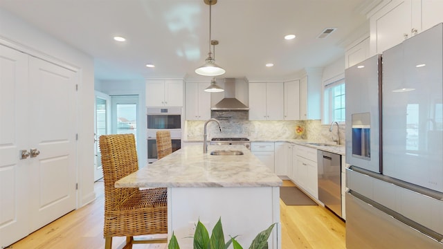 kitchen with stainless steel appliances, a kitchen island with sink, sink, wall chimney range hood, and white cabinets