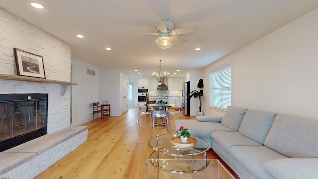 living room with ceiling fan with notable chandelier, light wood-type flooring, and a stone fireplace