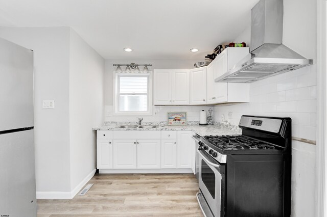 kitchen with white cabinets, light hardwood / wood-style flooring, stainless steel gas range, wall chimney exhaust hood, and white fridge