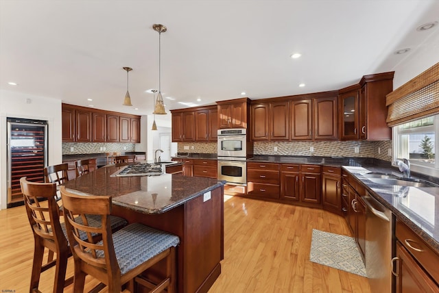 kitchen with light wood-type flooring, stainless steel appliances, sink, wine cooler, and hanging light fixtures