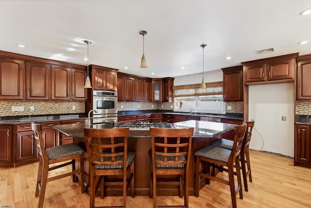 kitchen with appliances with stainless steel finishes, light wood-type flooring, a kitchen breakfast bar, pendant lighting, and an island with sink