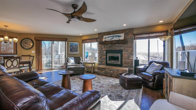 living room with dark wood-type flooring, ceiling fan with notable chandelier, and a stone fireplace