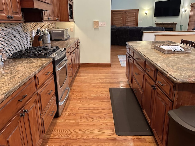 kitchen featuring wall chimney range hood, backsplash, light wood-type flooring, light stone counters, and stainless steel range with gas cooktop