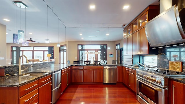 kitchen with wall chimney exhaust hood, decorative light fixtures, stainless steel appliances, dark stone counters, and sink