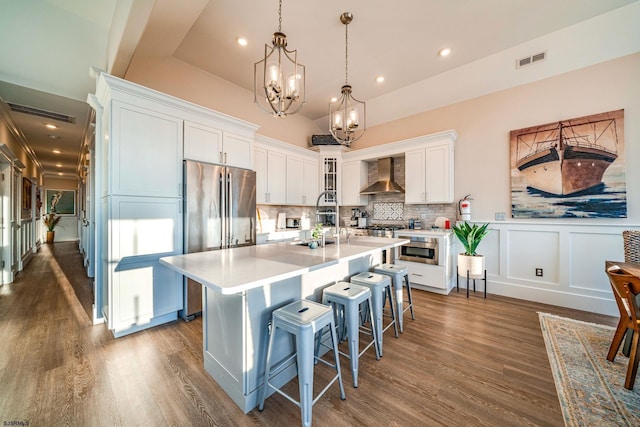 kitchen with pendant lighting, stainless steel fridge, white cabinets, a kitchen island with sink, and wall chimney range hood