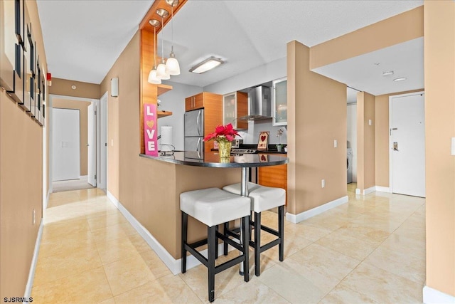 kitchen featuring wall chimney range hood, stainless steel refrigerator, hanging light fixtures, a kitchen bar, and kitchen peninsula