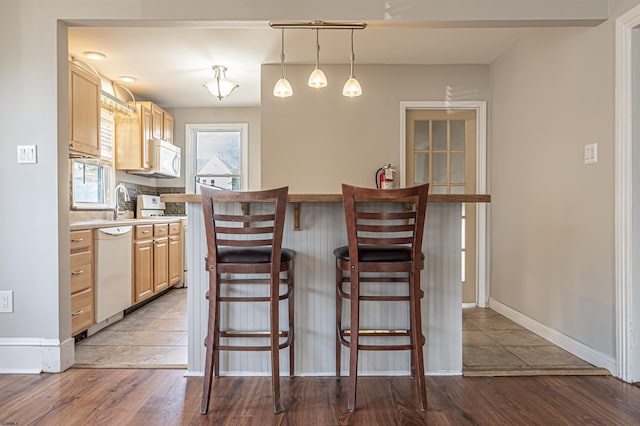 kitchen with light brown cabinetry, wood-type flooring, hanging light fixtures, a kitchen breakfast bar, and white appliances