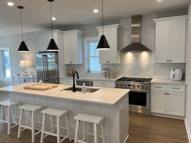 kitchen featuring white cabinetry, high quality appliances, an island with sink, pendant lighting, and wall chimney range hood