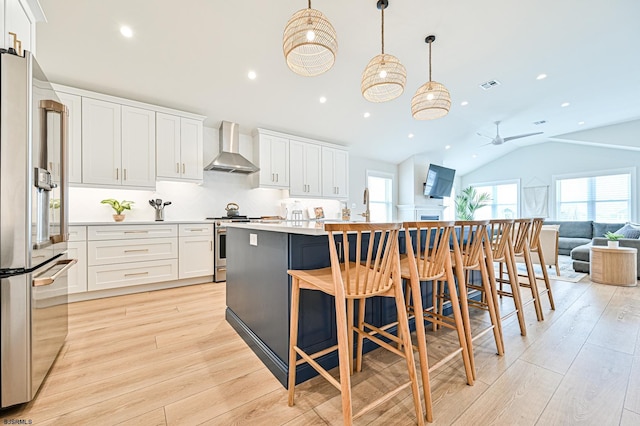 kitchen featuring appliances with stainless steel finishes, hanging light fixtures, white cabinets, a center island with sink, and wall chimney exhaust hood