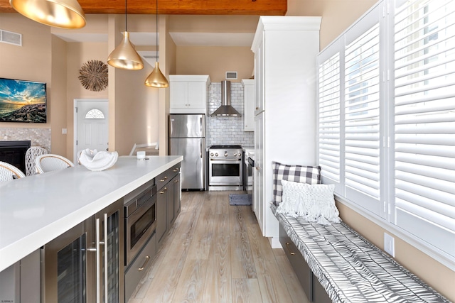 kitchen featuring appliances with stainless steel finishes, pendant lighting, white cabinetry, wall chimney range hood, and light hardwood / wood-style flooring