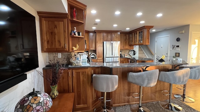 kitchen featuring stainless steel refrigerator, dark stone countertops, backsplash, a kitchen bar, and kitchen peninsula