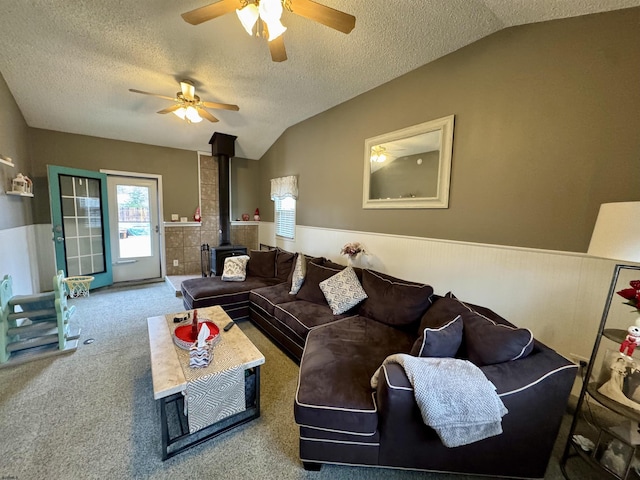 living room featuring vaulted ceiling, a wood stove, carpet, ceiling fan, and a textured ceiling