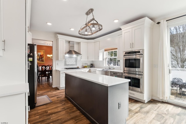 kitchen featuring wall chimney exhaust hood, white cabinetry, a center island, hanging light fixtures, and appliances with stainless steel finishes