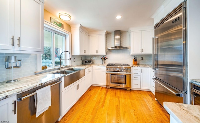 kitchen featuring light stone counters, a sink, high quality appliances, white cabinets, and wall chimney range hood