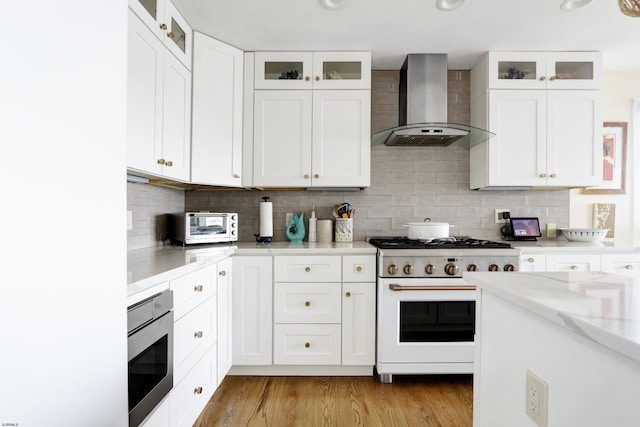 kitchen featuring wall chimney range hood, gas range oven, glass insert cabinets, and white cabinets