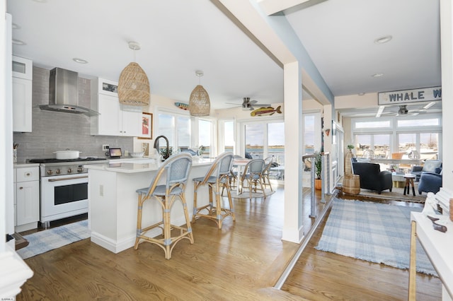 kitchen featuring open floor plan, hanging light fixtures, wall chimney range hood, white cabinetry, and gas range gas stove