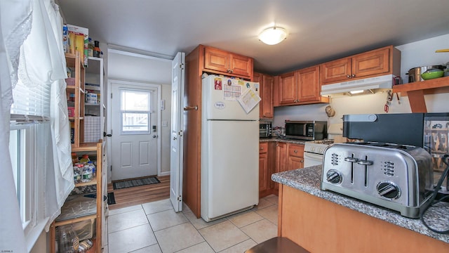 kitchen featuring light tile patterned flooring, under cabinet range hood, stainless steel microwave, freestanding refrigerator, and brown cabinetry