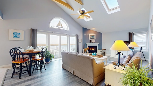 living area featuring a skylight, light wood finished floors, ceiling fan, high vaulted ceiling, and a warm lit fireplace