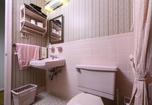 half bath featuring a wainscoted wall, tile walls, toilet, a sink, and wallpapered walls