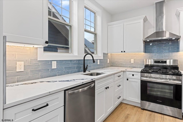 kitchen with white cabinets, light wood-style flooring, appliances with stainless steel finishes, wall chimney range hood, and a sink