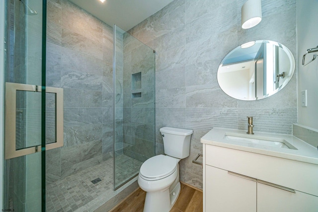 bathroom featuring toilet, wood finished floors, a tile shower, vanity, and tile walls