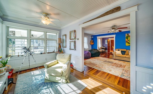 living area with wainscoting, ceiling fan, wood finished floors, crown molding, and a baseboard heating unit