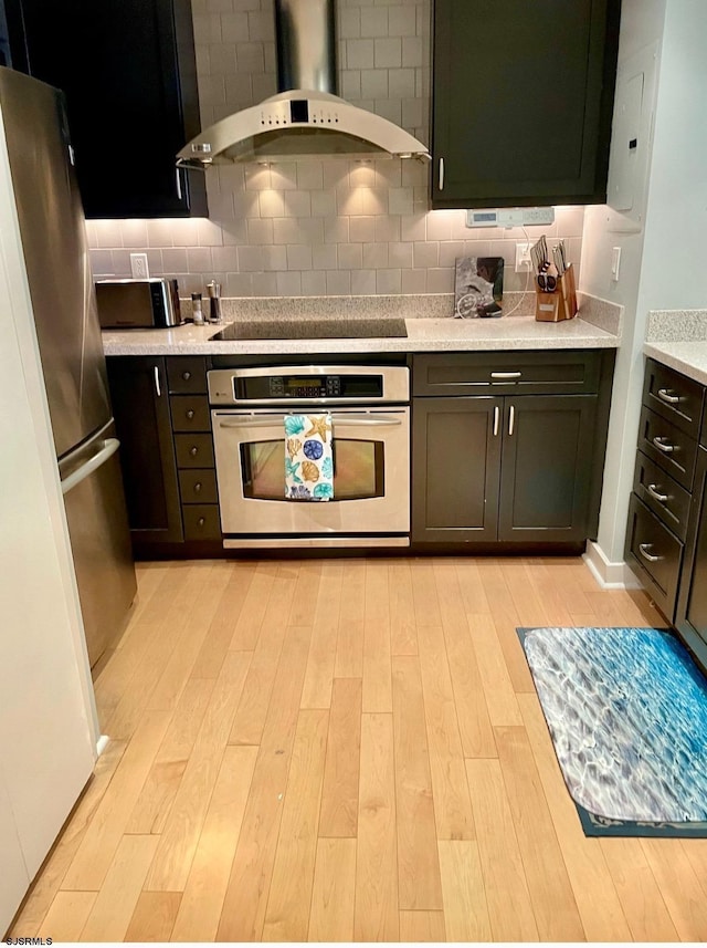 kitchen featuring light wood-style flooring, range hood, black electric stovetop, stainless steel oven, and backsplash