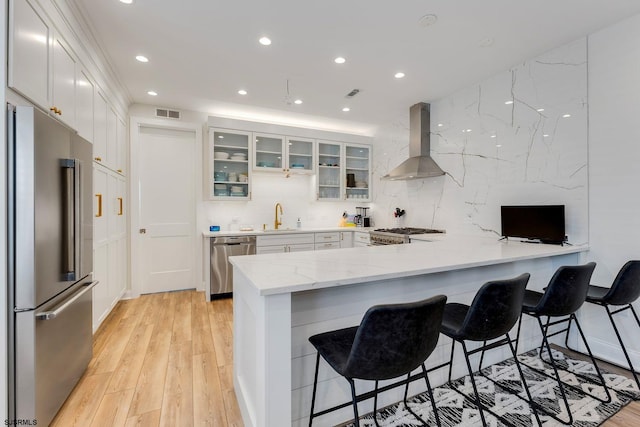 kitchen featuring glass insert cabinets, a peninsula, stainless steel appliances, wall chimney range hood, and a sink