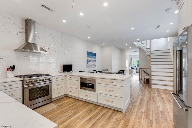 kitchen featuring a peninsula, visible vents, white cabinets, wall chimney range hood, and appliances with stainless steel finishes