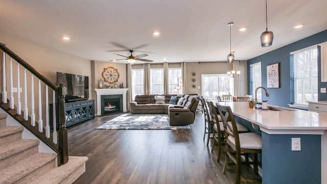 living area with a warm lit fireplace, recessed lighting, ceiling fan with notable chandelier, dark wood-type flooring, and stairway