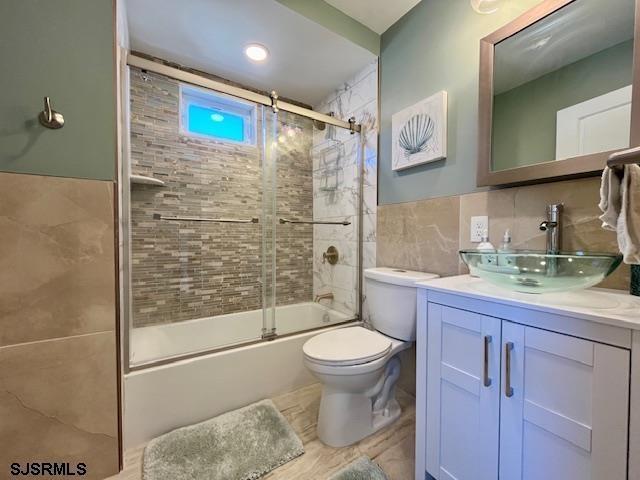 full bath with toilet, bath / shower combo with glass door, tile walls, and vanity