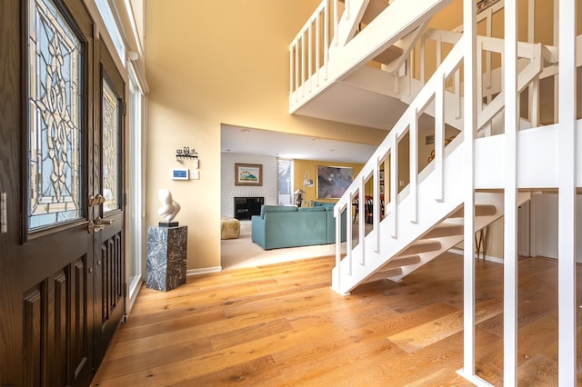 entrance foyer with stairway, light wood-style flooring, a towering ceiling, a brick fireplace, and baseboards