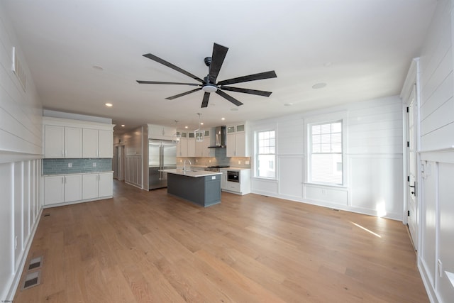 kitchen with built in refrigerator, a kitchen island with sink, light wood-type flooring, wall chimney range hood, and backsplash