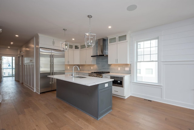 kitchen with built in refrigerator, wall chimney range hood, light wood-style flooring, and a sink