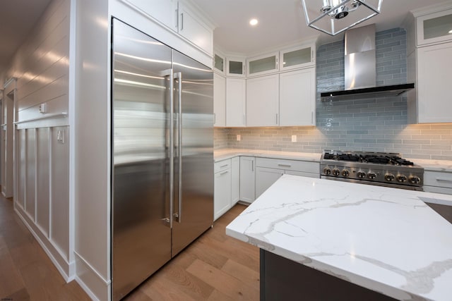 kitchen featuring light wood-style flooring, white cabinets, stainless steel built in fridge, wall chimney range hood, and range