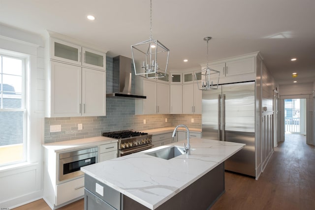 kitchen featuring backsplash, a kitchen island with sink, a sink, wall chimney range hood, and built in appliances