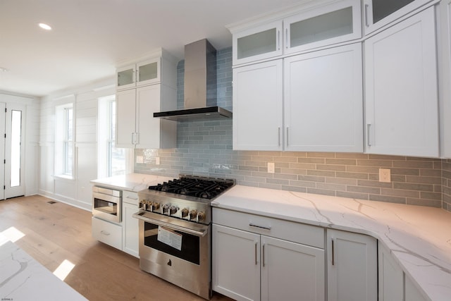 kitchen featuring stainless steel range, wall chimney exhaust hood, light wood-style flooring, white cabinetry, and backsplash