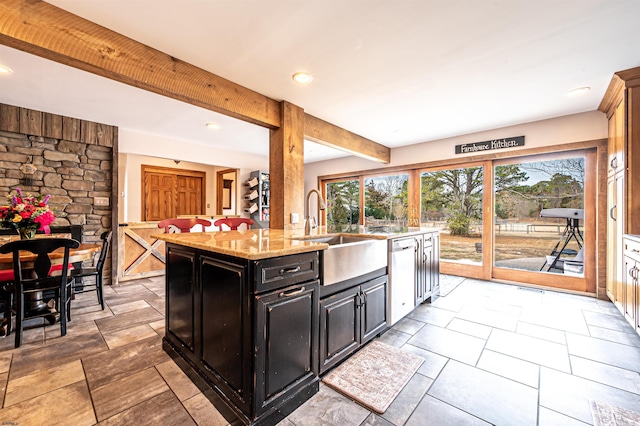 kitchen with light stone counters, dark cabinets, a sink, beamed ceiling, and a center island with sink