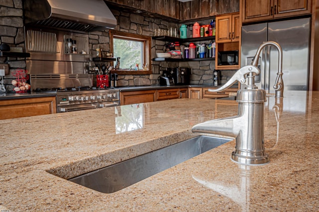 kitchen featuring light stone counters, brown cabinets, stainless steel appliances, wall chimney range hood, and open shelves