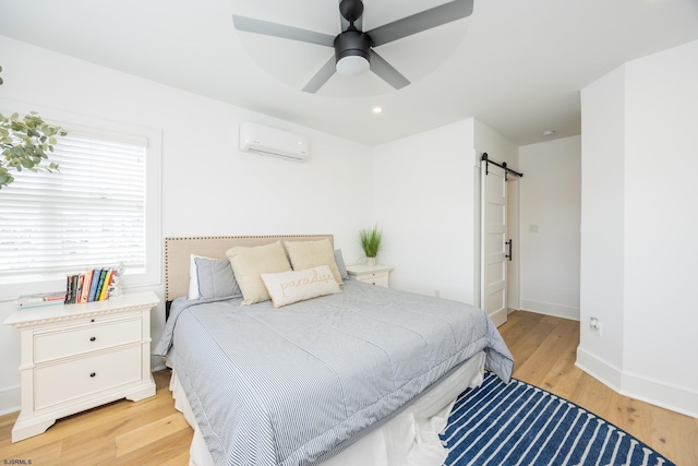 bedroom featuring ceiling fan, a barn door, baseboards, light wood-style floors, and a wall mounted air conditioner