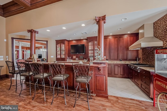 kitchen with wall chimney range hood, visible vents, decorative columns, a kitchen breakfast bar, and tasteful backsplash