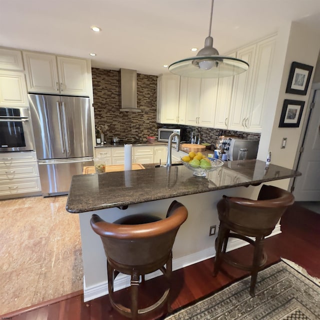 kitchen featuring dark stone counters, appliances with stainless steel finishes, a peninsula, wall chimney range hood, and decorative backsplash