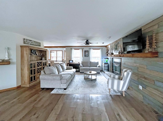 living area with a glass covered fireplace, ceiling fan, baseboards, and wood finished floors