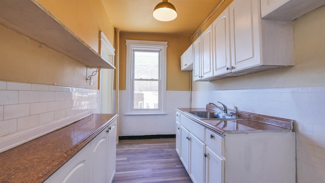 kitchen with a sink, dark stone countertops, wood finished floors, white cabinetry, and wainscoting