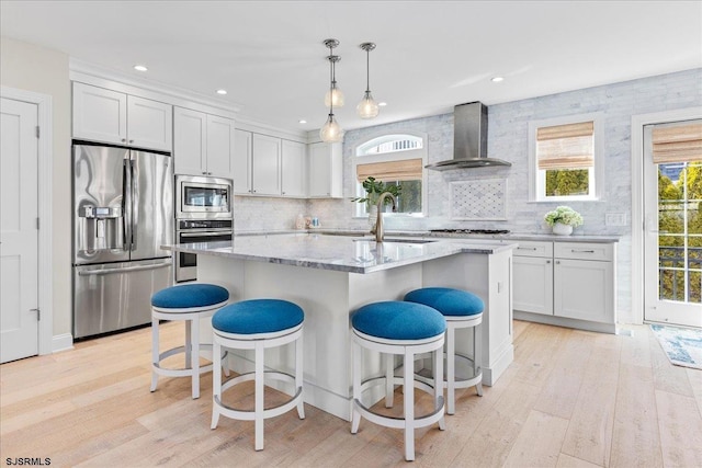 kitchen with wall chimney range hood, light stone countertops, an island with sink, stainless steel appliances, and a sink