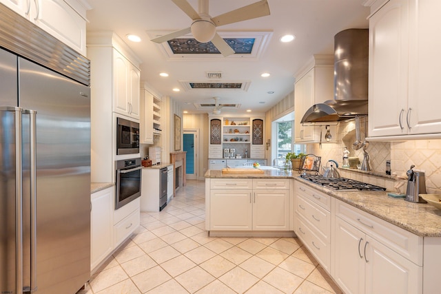 kitchen featuring a peninsula, light tile patterned flooring, white cabinets, built in appliances, and wall chimney range hood