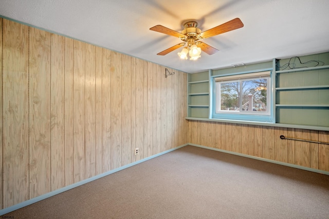 empty room with baseboards, carpet, ceiling fan, built in features, and wood walls