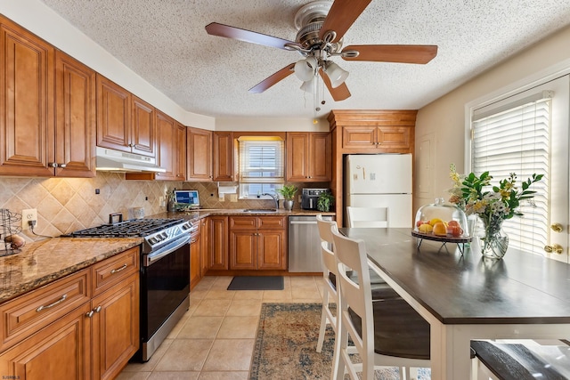 kitchen with ceiling fan, under cabinet range hood, appliances with stainless steel finishes, brown cabinetry, and a sink