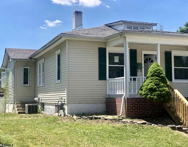 view of side of home with a shingled roof, central air condition unit, covered porch, a chimney, and a yard